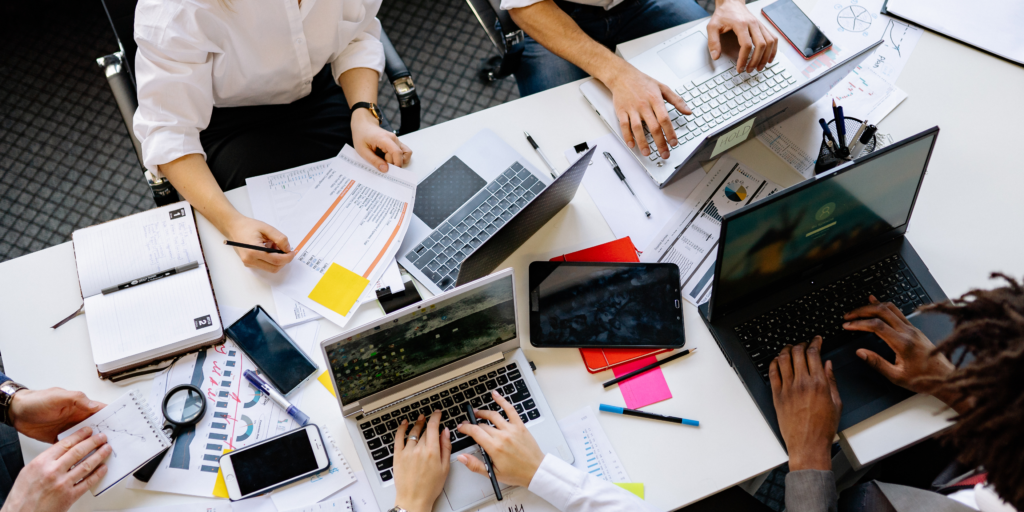 An overhead shot shows several people working around a white table. The table is covered with laptops, smartphones, a tablet, notebooks, pens, and printed charts. Each person is actively engaged with their devices or paperwork, suggesting a collaborative and busy work session.