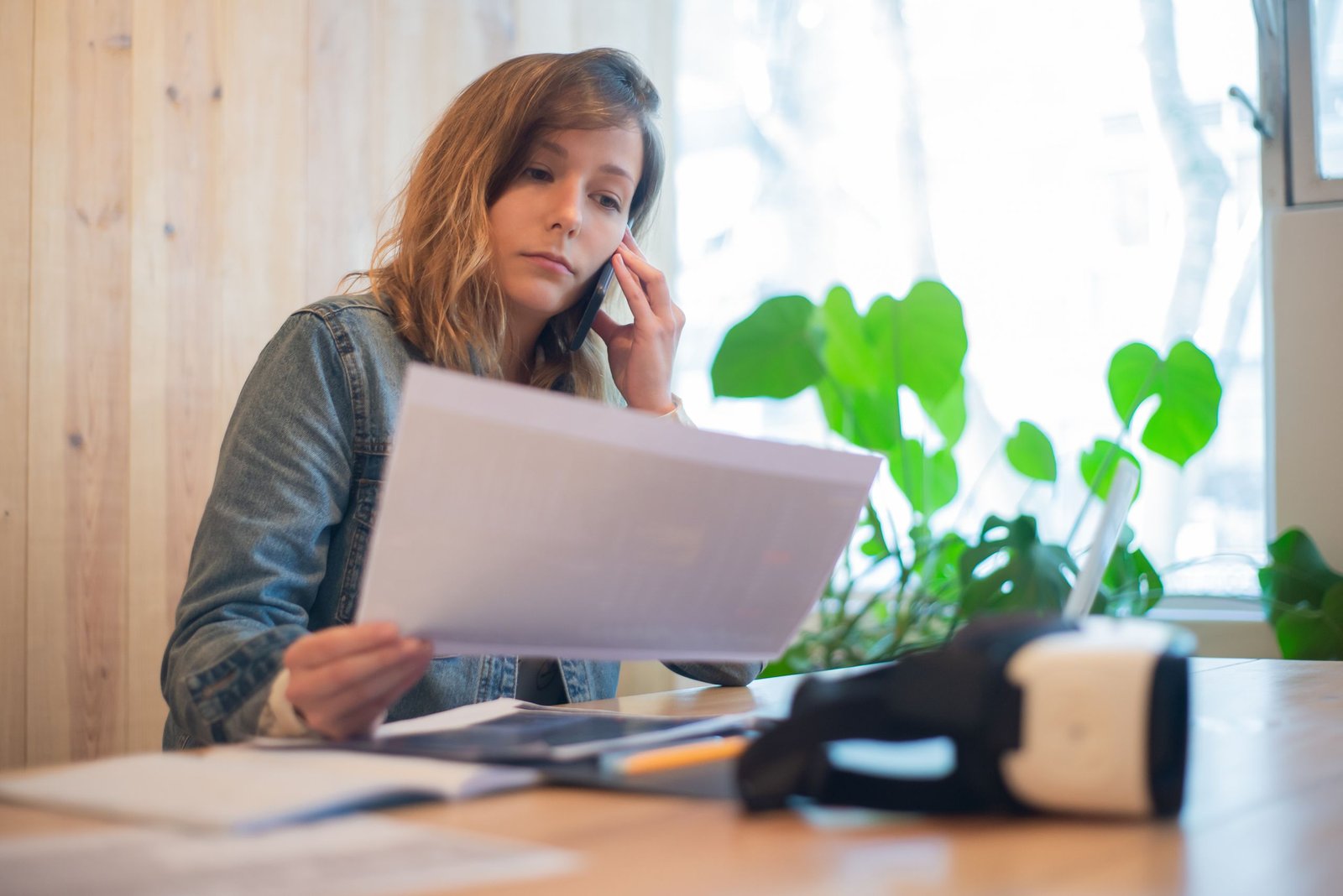 Businesswoman reviewing paperwork while on a call at a modern office workspace.